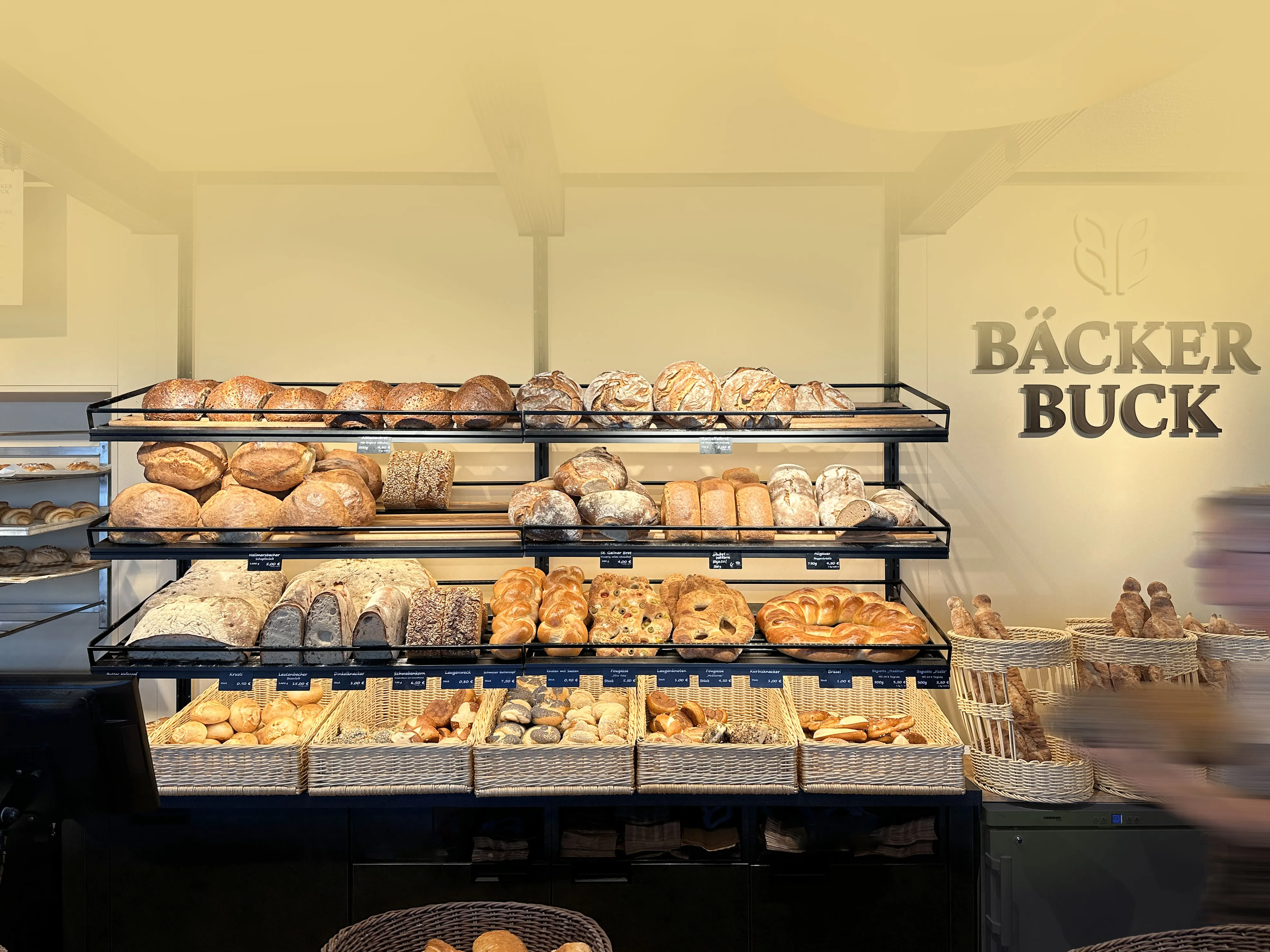 Brotregal in der Bäckerei Buck mit Laiben, Zöpfen und Brezeln, daneben der Schriftzug Bäcker Buck an der Wand